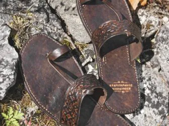 Dark brown leather sandals on grey stones, surrounded by some grass and plants.