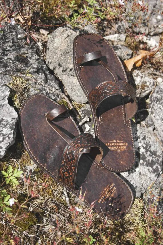 Dark brown leather sandals on grey stones, surrounded by some grass and plants.