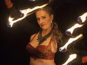 This photo shows the leather costume in full seen a little bit from a side. Costume is worn by the artist and she holds two sets of burning torches up in top left and down right corners of the photo.