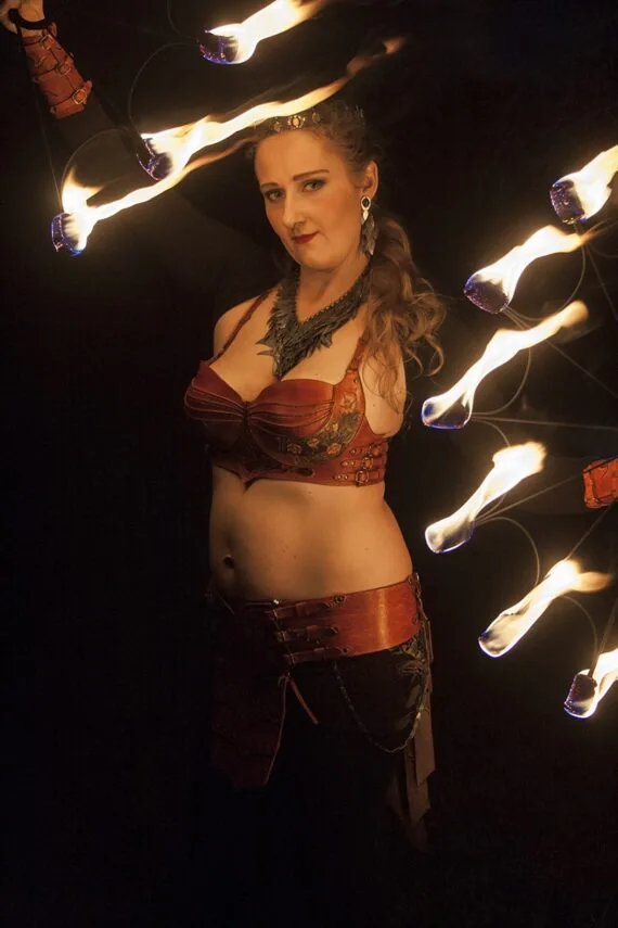 This photo shows the leather costume in full seen a little bit from a side. Costume is worn by the artist and she holds two sets of burning torches up in top left and down right corners of the photo.