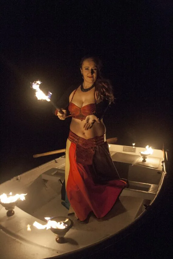 This is a photo featuring the leather costume for the fire artist. Photo is taken on the lake shore. Artist stands in the boat and there are goblets of fire on the boat edges. Artist holds a big burning torch in one hand and reaches the other towards the viewer.