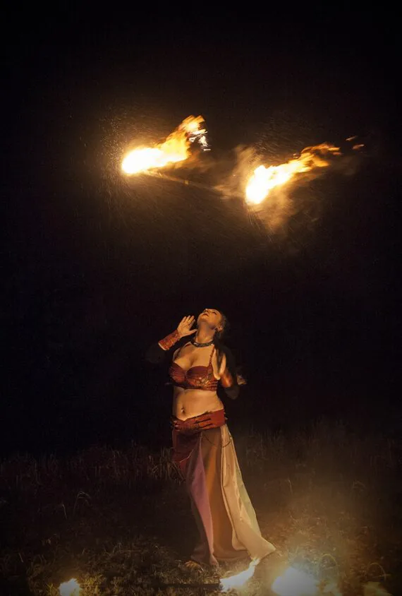 Photo shows the leather costume for a fire artist. Photo is taken in darker time. Artist has just thrown the staff with burning ends into the air above her head and is looking up towards it.