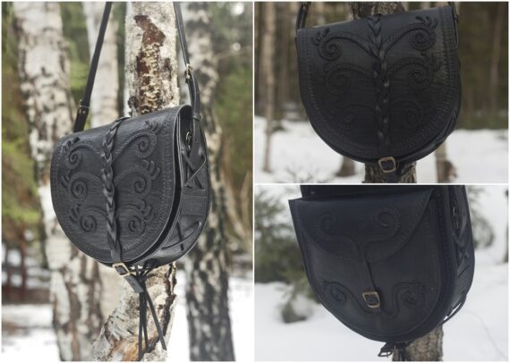 Black round shaped leather shoulder bag that is decorated by hand carved image and on sewn leather laces. Laces are wet-formed to be curvy and make up a decorative pattern. Photo is taken in a snowy forest. Bag hangs on the birch tree branch and there are some evergreen trees visible on the background.