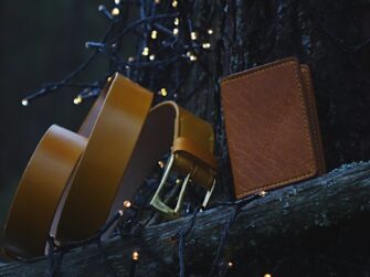 Photo is taken in the darkening forest where the belt and a card wallet is set onto the fallen tree. The whole scene surrounding the yellow leather goods is a bit blue toned. Some small sparkling lights in the background and foreground give it a nice warmer feeling.