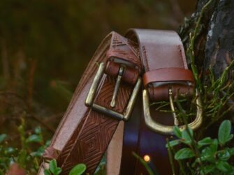 Two belts are set to lean to the tree trunk. Belts are surrounded by forest plants and there is a deep darker green background which stems from the actual forest trees and plants farther away.