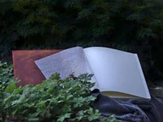 Square shaped album is opened and placed onto the woolen blanket on the stone wall. Album is surrounded by rather darker green bushes.