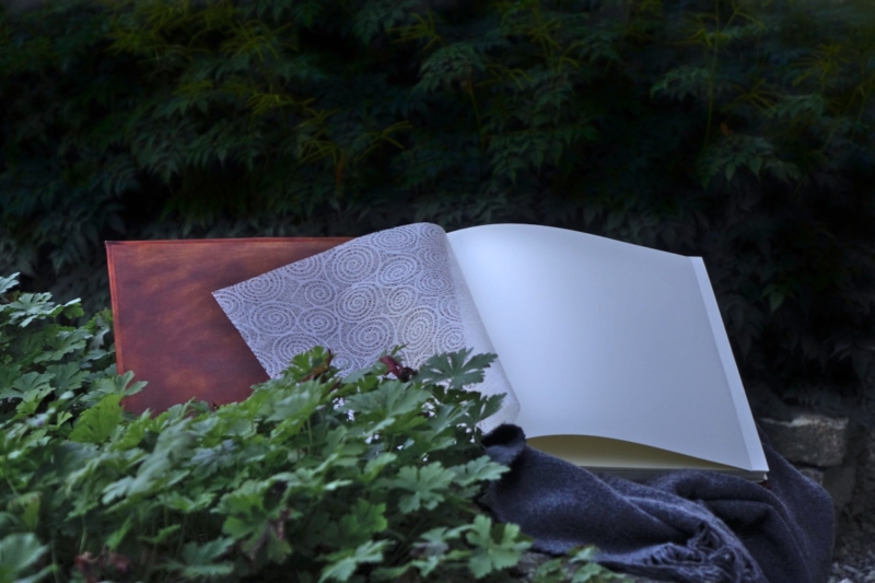 Square shaped album is opened and placed onto the woolen blanket on the stone wall. Album is surrounded by rather darker green bushes.