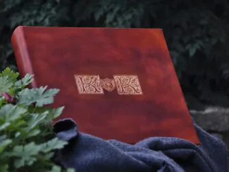 Square shaped album is placed onto the woolen blanket on the stone wall. Album is surrounded by rather darker green bushes.