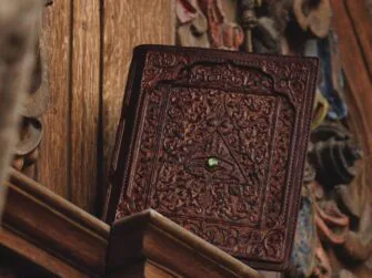 Leather covers displayed on the wooden panel. Panel is carved and painted in multiple colors. this photo features the front side of the covers that is decorated also by a small green precious stone.