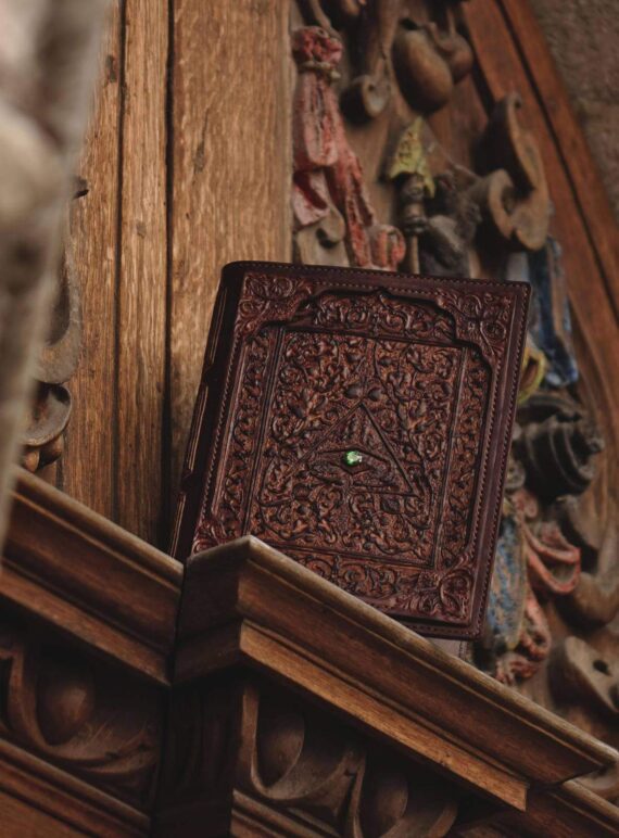 Leather covers displayed on the wooden panel. Panel is carved and painted in multiple colors. this photo features the front side of the covers that is decorated also by a small green precious stone.