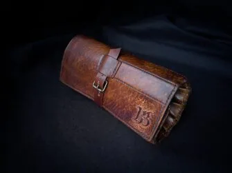 Brown leather tools roll photographed on the black canvas background. The roll is rolled up and fastened.