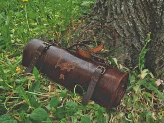 Knife roll is placed on the grass besides tree roots. Roll is closed and fastened by two straps which may be opened by small buckles.