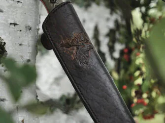 Knife sheath in on the white birch branch, red and green colored rowan branches on the background. The focus of this photo is on the front side of the sheath.