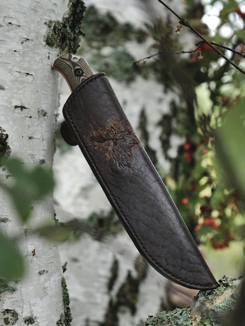 Knife sheath in on the white birch branch, red and green colored rowan branches on the background. The focus of this photo is on the front side of the sheath.