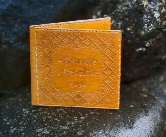 Photo shows a decorated yellow leather covers for a cd record. Covers are placed upright onto wet dark large stones. There are hand carved decorations on the cover - a pattern and text "To beloved Mirtel 2016".