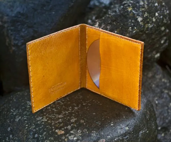 Photo shows a decorated yellow leather covers for a cd record. Covers are placed upright onto wet dark large stones and are opened towards the viewer. There is a leather pocket with a cd in it on the right side of the covers.