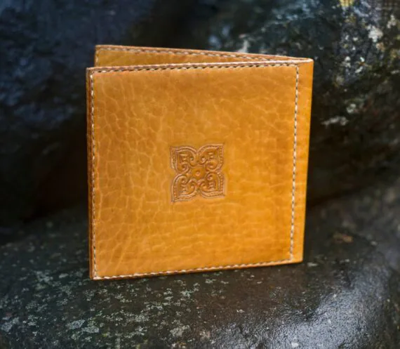 Photo shows a decorated yellow leather covers for a cd record, featuring the back side of the covers. There is a small curvy carved motif in the centre of the leather covers. Covers are placed upright onto wet dark large stones.