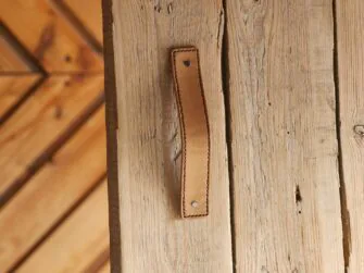 Indoor photo of the leather handle attached to the wooden door. Light brown handle with stitched edges.