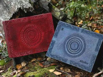 Large leather boxes in mahogany and blue are set onto the forest ground by the fallen and broken tree trunk. There are leaves all over the ground and some moss is visible between the tree roots. Mahogany box has carving of a sun-like interpretation and blue features a moon themed decoration on the lid. There is also some folk pattern carved around the edges, a name on the right bottom corner and Roman numbering on the left top corner.