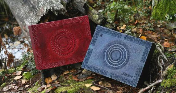 Large leather boxes in mahogany and blue are set onto the forest ground by the fallen and broken tree trunk. There are leaves all over the ground and some moss is visible between the tree roots. Mahogany box has carving of a sun-like interpretation and blue features a moon themed decoration on the lid. There is also some folk pattern carved around the edges, a name on the right bottom corner and Roman numbering on the left top corner.