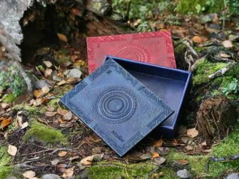 Large boxes in mahogany and blue are set onto the forest ground by the fallen and broken tree trunk. Blue box is placed to the front of the mahogany box and the lid is slightly pushed aside. It is visible that the box is covered by leather also on the inside. There are leaves all over the ground and some moss is visible between the tree roots.