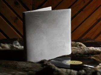 White leather covers for vinyl record, photographed indoors. Covers are placed onto hairy skin and there is a wooden wall further in the back. Vinyl record is laid down besides the covers. Covers are set upright, slightly opened and facing the viewer. There is a small inscription on the front cover.