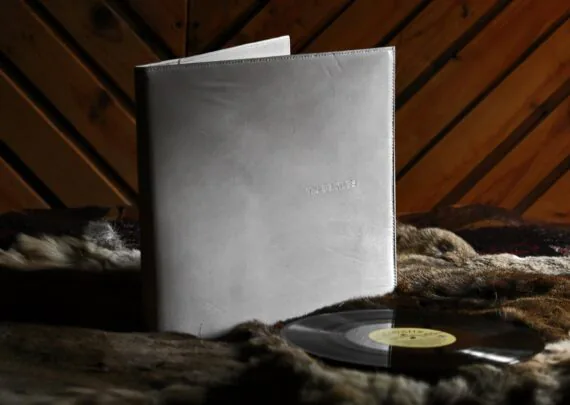 White leather covers for vinyl record, photographed indoors. Covers are placed onto hairy skin and there is a wooden wall further in the back. Vinyl record is laid down besides the covers. Covers are set upright, slightly opened and facing the viewer. There is a small inscription on the front cover.