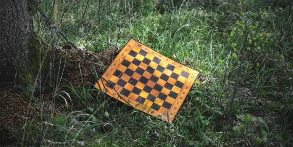 Leather chess board is placed onto the grass, near to the tree. The chess board is made in yellow and dark brown.