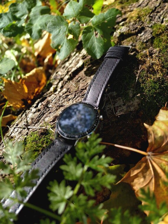 Watch is placed on the mossy the tree root, surrounded by autumn leaves in yellow and green.